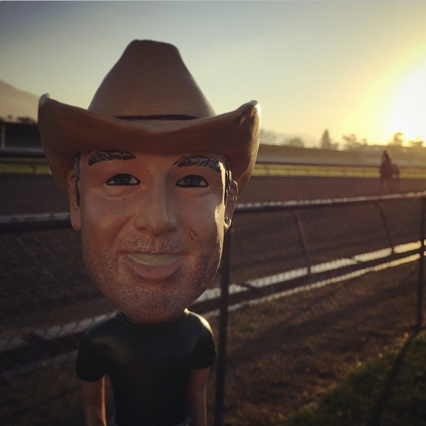 Mountain Man Steve Bobblehead in a cowboy hat, set against a sunset backdrop at a racetrack, highlighting outdoor cooking themes.
