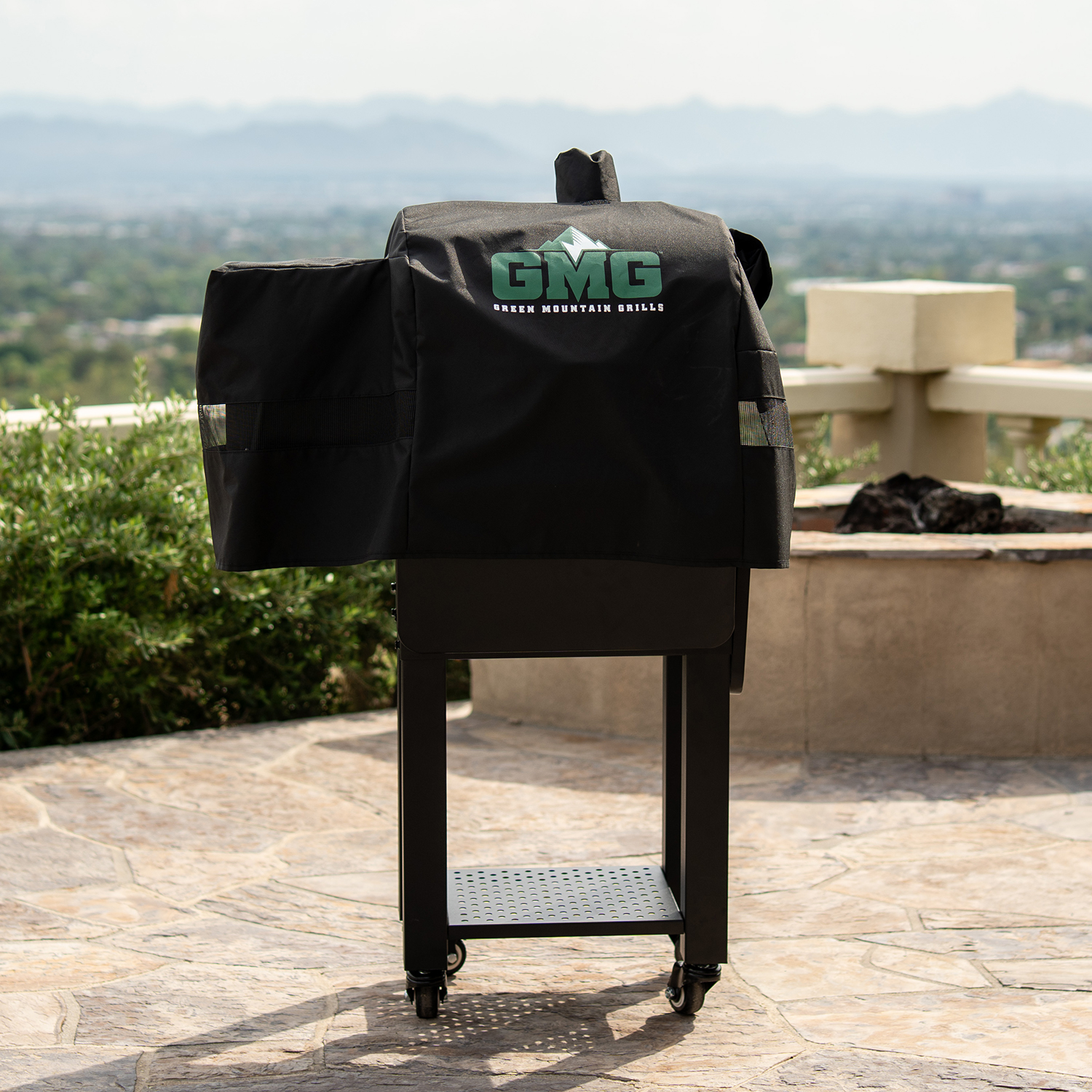 Portable smoker grill covered with a black protective cover, featuring the Green Mountain Grills logo, set against a scenic outdoor backdrop with mountains and greenery, emphasizing its compact design for outdoor cooking.